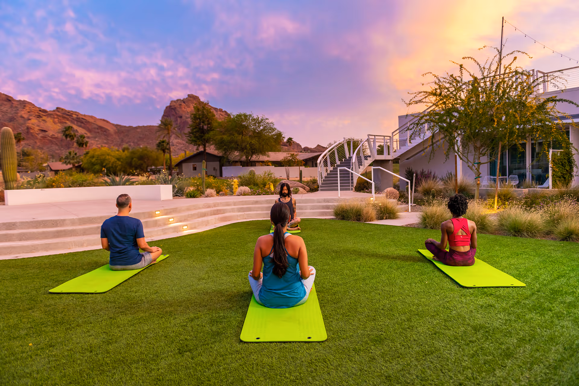 Three people sitting cross-legged on yoga mats meditating outdoors on green grass at sunset with mountains and resort buildings in the background.